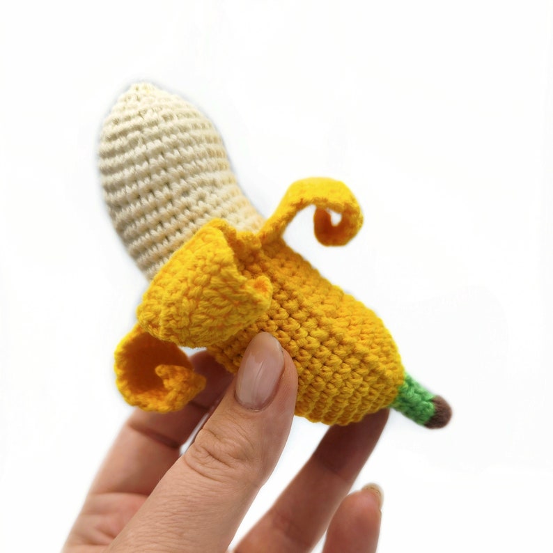 May include: A crocheted banana toy with a yellow peel and a cream-colored top. The banana is held in a hand.
