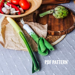May include: A crochet pattern for a green leek with white accents. The pattern is shown on a table with a real leek, artichoke, and red peppers.