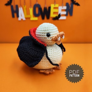 May include: A crocheted amigurumi duck wearing a black cape with a red lining and fangs. The duck is sitting on an orange surface with a wooden sign that says "HALLOWEEN" hanging above it. The sign is decorated with bats.