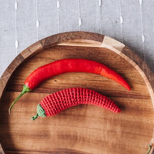 Puede incluir: Dos chiles rojos en una bandeja de madera. Un chile es real y el otro est&aacute; tejido a crochet.