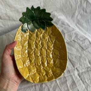 May include: A ceramic dish shaped like a pineapple with a yellow textured body and a dark green leafy top. The surface of the dish features a raised, grid-like pattern mimicking the skin of a pineapple, finished with a glossy glaze throughout.