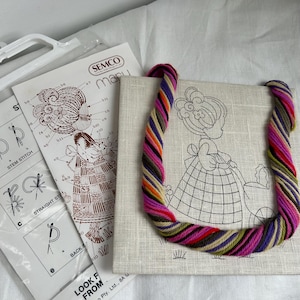 Puede incluir: Un kit de bordado Semco que incluye un panel de tela cuadrado con el contorno impreso de una persona empujando un cochecito, acompañado de una madeja de hilo multicolor en tonos rosa, morado, amarillo y verde, y una guía de instrucciones.