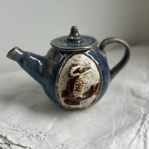 May include: A ceramic teapot with a glossy blue glaze and a dark handle. The centre features a raised, circular relief panel with a speckled cream background depicting a brown and white kookaburra bird perched on a branch with a yellow beak.