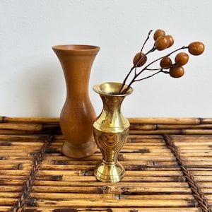 May include: A smooth, light brown wooden vase with a flared rim and a rounded body stands next to a polished gold-tone brass vase with a flared rim and a slender neck. A thin, dark brown branch with several small, round, light brown berries is inside.