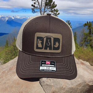 May include: Brown and tan trucker hat with a brown leather patch that says "DAD" with a fishing hook and a fish silhouette. The hat has a mesh back and the words "Richardson 112 Original Trucker Adjustable" on the back.