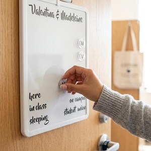 May include: A white magnetic dry-erase board with a black frame, featuring the text "Valentina & Madeleine" at the top and a list of locations including "here," "in class," "sleeping," "on campus," and "student union." A black marker is attached.