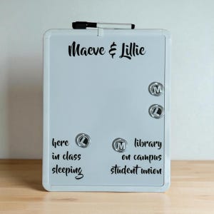 May include: A white rectangular magnetic dry erase board with a black marker attached to the top. The board features the black text Maeve & Lillie at the top, with additional text below and four circular magnets labeled with the letters M and L.