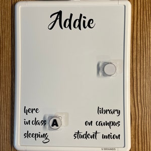 May include: A white dry erase board with the name Addie printed at the top. The board features text options including here, in class, sleeping, library, on campus, and student union. A black dry erase marker is attached to the top frame.