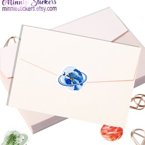 May include: A stack of pink envelopes with a blue and white watercolour sticker of the Earth with a ring around it. The sticker is on the top envelope. There are two other stickers, one green and one red, visible in the image.