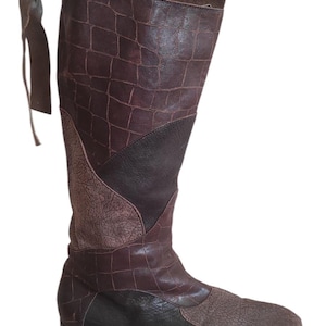 以下が含まれることがあります： 茶色の膝丈ブーツ。革製で、シャフトにはクロコダイル風の模様があります。ブーツは折り返しのあるトップと、さまざまな質感と茶色の濃淡を組み合わせており、フラットソールです。