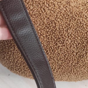 May include: A brown, textured handbag with a dark brown leather strap. The bag's body has a plush, fuzzy appearance. The strap is smooth and has a subtle, pebbled texture. The bag is a crescent shape.