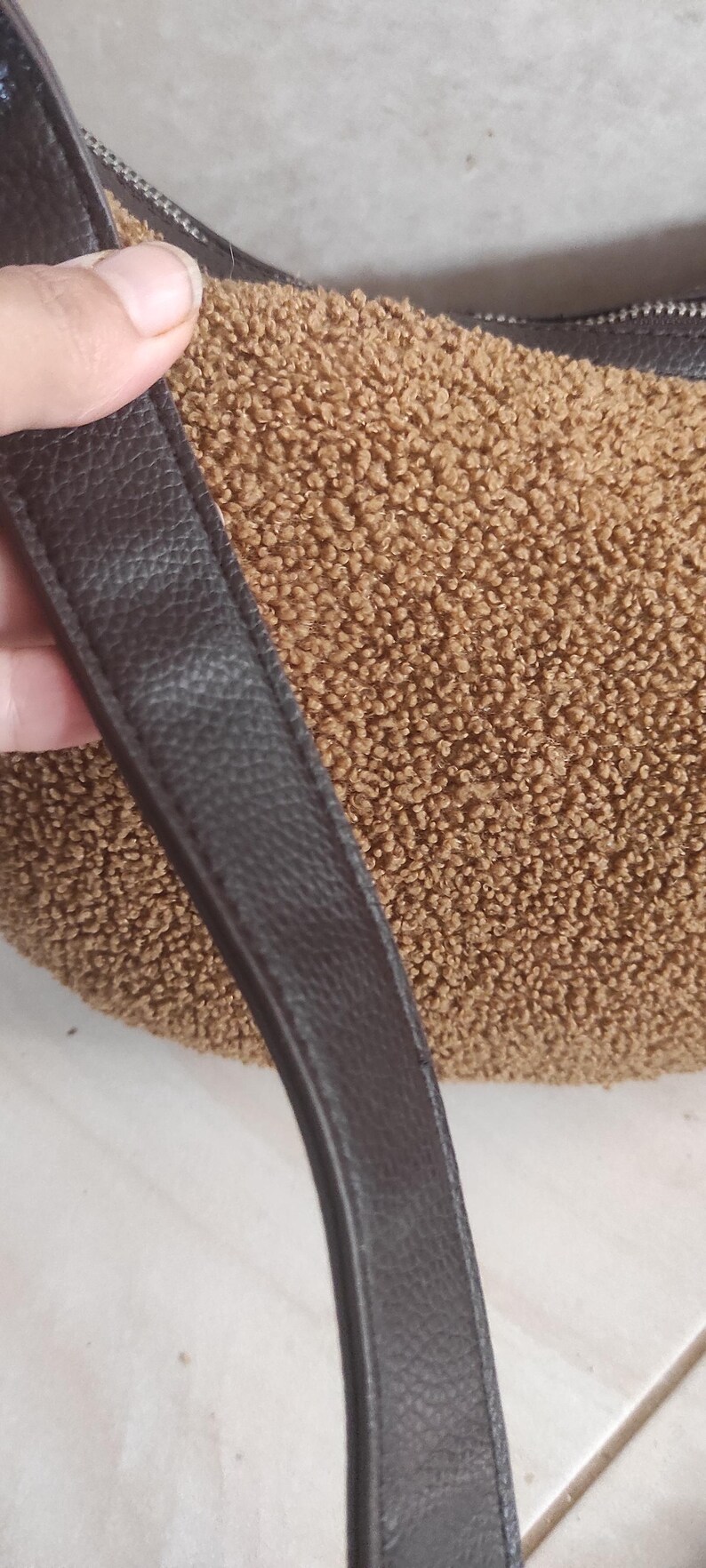 May include: A brown, textured handbag with a dark brown leather strap. The bag's body has a plush, fuzzy appearance. The strap is smooth and has a subtle, pebbled texture. The bag is a crescent shape.
