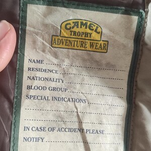 May include: A tan and green label with the text "Camel Trophy Adventure Wear" and a form for name, residence, nationality, blood group, special indications, and in case of accident please notify.