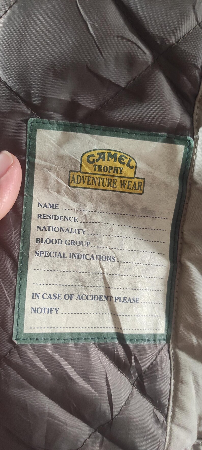 May include: A tan and green label with the text "Camel Trophy Adventure Wear" and a form for name, residence, nationality, blood group, special indications, and in case of accident please notify.