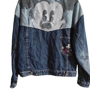 以下が含まれることがあります： 背面にミッキーマウスのグラフィックが施された青いデニムジャケット。ジャケットの左側に、より小さなミッキーマウスのグラフィックが描かれたポケットがあります。