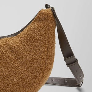 May include: A brown, crescent-shaped shoulder bag with a textured, fuzzy exterior. The bag features a dark brown leather strap and trim. The strap is adjustable and attaches to the bag with metal hardware. The bag is a neutral color.
