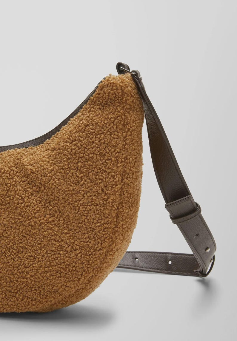 May include: A brown, crescent-shaped shoulder bag with a textured, fuzzy exterior. The bag features a dark brown leather strap and trim. The strap is adjustable and attaches to the bag with metal hardware. The bag is a neutral color.