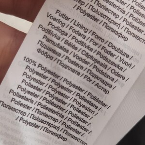 May include: A close-up of a white fabric tag with text in multiple languages. The tag lists the material composition as 100% polyester and includes terms like "Futter", "Voering", and "Pod&scaron;&iacute;vka".