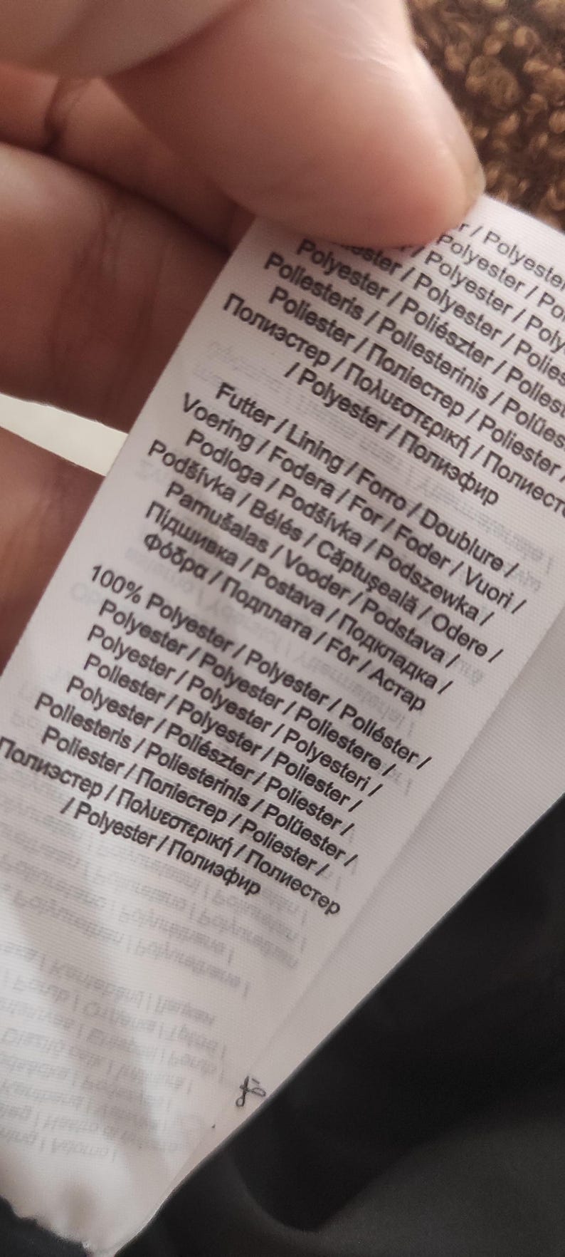 May include: A close-up of a white fabric tag with text in multiple languages. The tag lists the material composition as 100% polyester and includes terms like "Futter", "Voering", and "Pod&scaron;&iacute;vka".