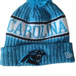 May include: A blue knit beanie with a matching pom-pom. The word "CAROLINA" is displayed in white block letters across the front. A black panther logo is on the front, and a New Era logo is on the side.