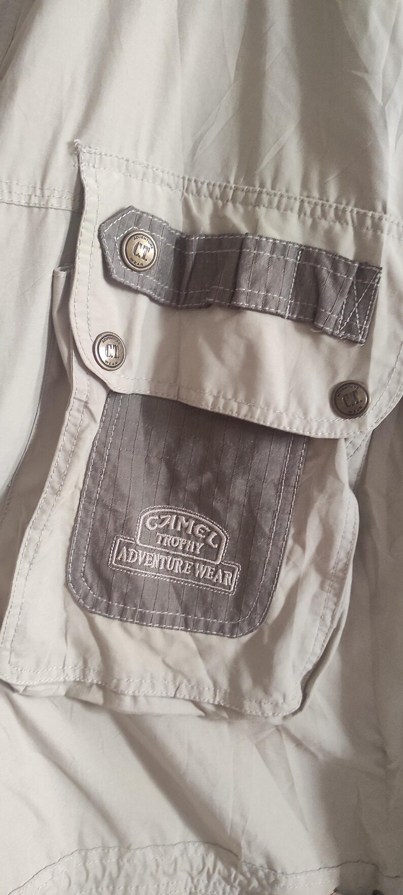 May include: A close-up of a gray and beige jacket with a pocket featuring the text "C.T. Trophy Adventure Wear".