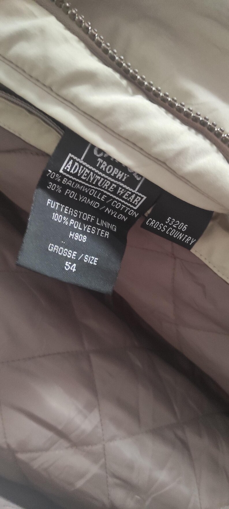 May include: A black label with white text that reads "Adventure Wear Trophy" and "Cross Country 53206". The label also includes details about the fabric composition: 70% cotton, 30% nylon, and a lining made of 100% polyester. The size is indicated as "54".