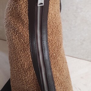 May include: A brown, textured crossbody bag with a silver zipper and black leather trim. The bag has a fluffy, teddy bear-like texture. The zipper pull has a logo. The strap is black.