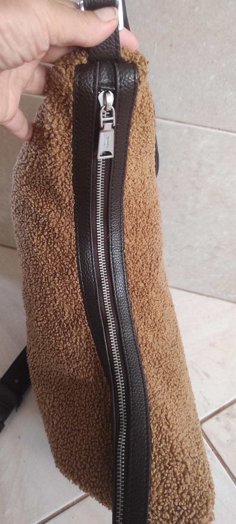 May include: A brown, textured crossbody bag with a silver zipper and black leather trim. The bag has a fluffy, teddy bear-like texture. The zipper pull has a logo. The strap is black.