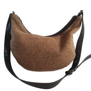 May include: A brown, textured shoulder bag with a curved shape. The bag features a black zipper and a black adjustable strap. The bag's material has a soft, plush appearance. The strap is attached to the bag with black hardware.