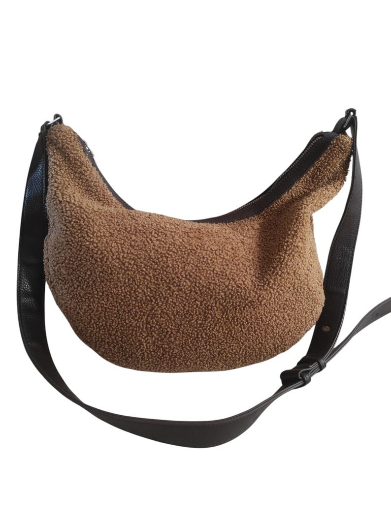May include: A brown, textured shoulder bag with a curved shape. The bag features a black zipper and a black adjustable strap. The bag's material has a soft, plush appearance. The strap is attached to the bag with black hardware.