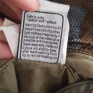 May include: A white fabric care label with black text that reads "Fabric with "WASH OUT" effect. Warning: Dark colors could bleed (lose color) to other light clothes. Irregularity in the look of the material is possible and on purpose. Wash separately. Inside out. Colors will slightly fade after each washing. Don't try to remove single spots."