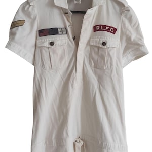 Puede incluir: Vestido crema de manga corta con cuello y cierre de botones. Cuenta con dos bolsillos en el pecho y parches con texto y símbolos. Incluye cintura con cordón y charreteras.