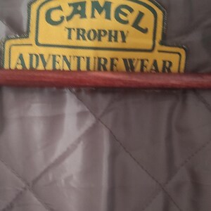 May include: A close-up of a gray quilted jacket with a yellow and green patch that reads "Camel Trophy Adventure Wear".