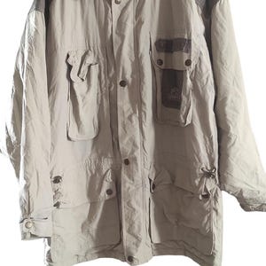 May include: A beige hooded jacket with brown accents and multiple pockets. The jacket has a button closure and a drawstring at the waist.
