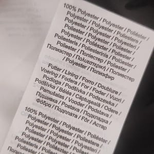 May include: A close-up of a white fabric tag with text in multiple languages. The tag lists the material as 100% polyester and includes information about lining and other details. The tag is partially visible.