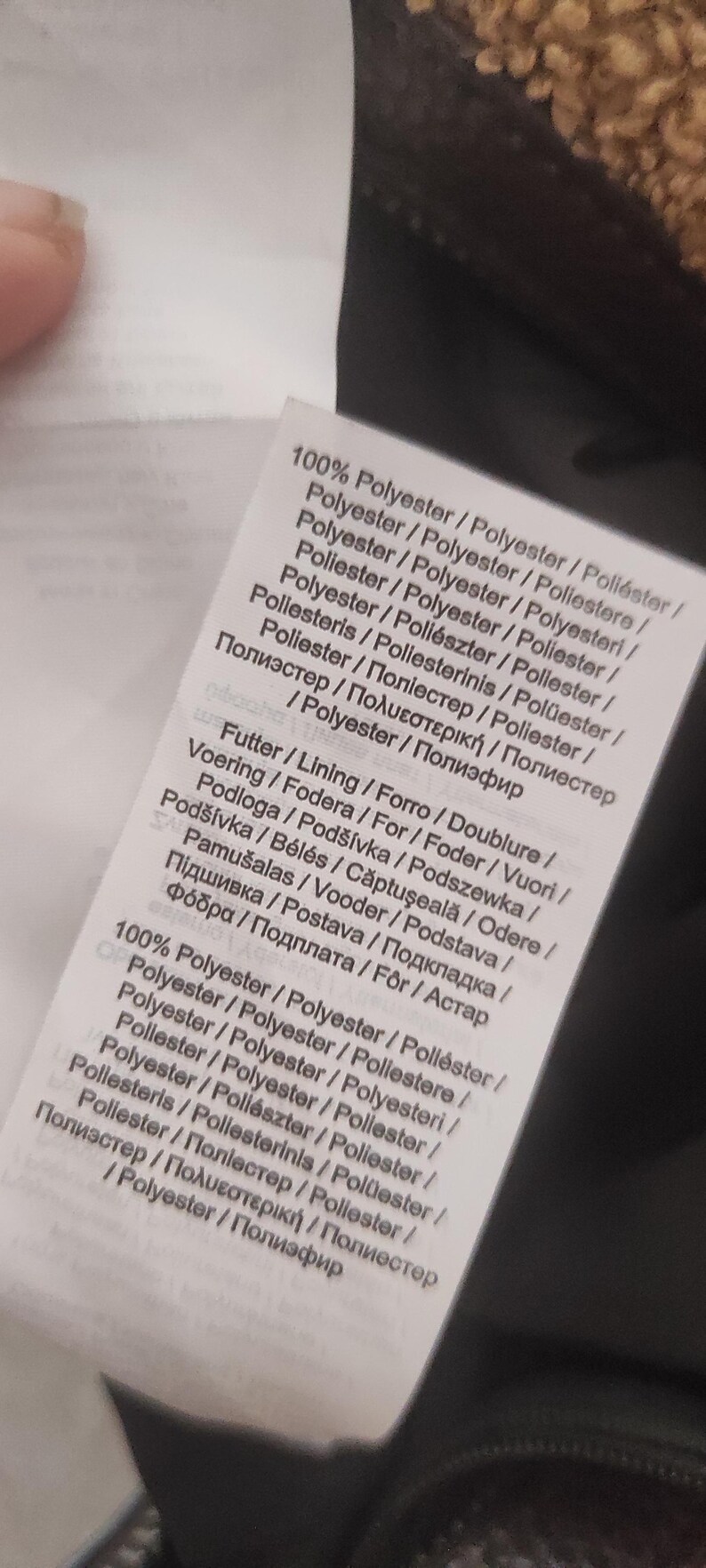 May include: A close-up of a white fabric tag with text in multiple languages. The tag lists the material as 100% polyester and includes information about lining and other details. The tag is partially visible.