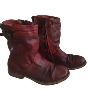 May include: A pair of distressed red leather boots. The boots have a mid-calf height, with a side zipper and buckle details. The leather has a worn, vintage appearance, with visible creases and variations in color.