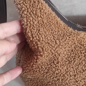 May include: A brown, fluffy shoulder bag with a black strap and zipper. The bag has a soft, textured exterior, and a black zipper. The bag is a neutral color and can be used for many occasions.