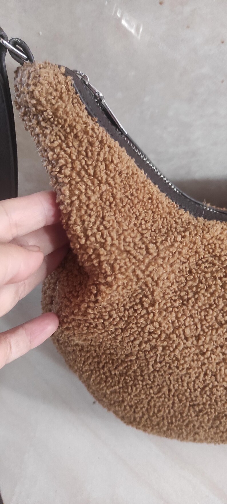 May include: A brown, fluffy shoulder bag with a black strap and zipper. The bag has a soft, textured exterior, and a black zipper. The bag is a neutral color and can be used for many occasions.