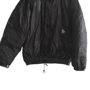 Puede incluir: Chaqueta acolchada negra con cuello alto y un pequeño logotipo triangular en el pecho. La chaqueta tiene cierre de cremallera y un cordón en la parte inferior. La chaqueta está colgada en una percha de madera.