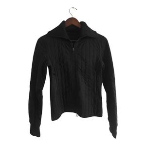 Puede incluir: Una chaqueta negra acolchada con cierre de cremallera completa y cuello ancho. La chaqueta tiene mangas largas y dos bolsillos laterales. Cuelga de una percha de madera sobre un fondo blanco.