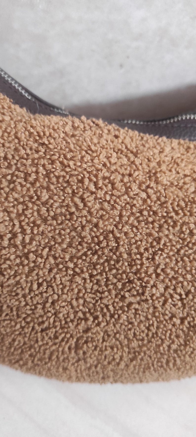 May include: Close-up of a brown, textured handbag. The bag has a soft, fuzzy exterior and a dark brown strap. The bag's material has a plush, teddy bear-like appearance.