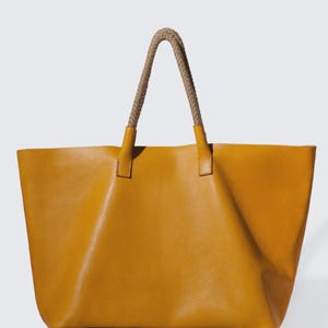 May include: Mustard yellow tote bag with rope handles. The bag is made of a smooth material and has a rectangular shape. The handles are a light brown color and are attached to the bag with small, matching yellow tabs.
