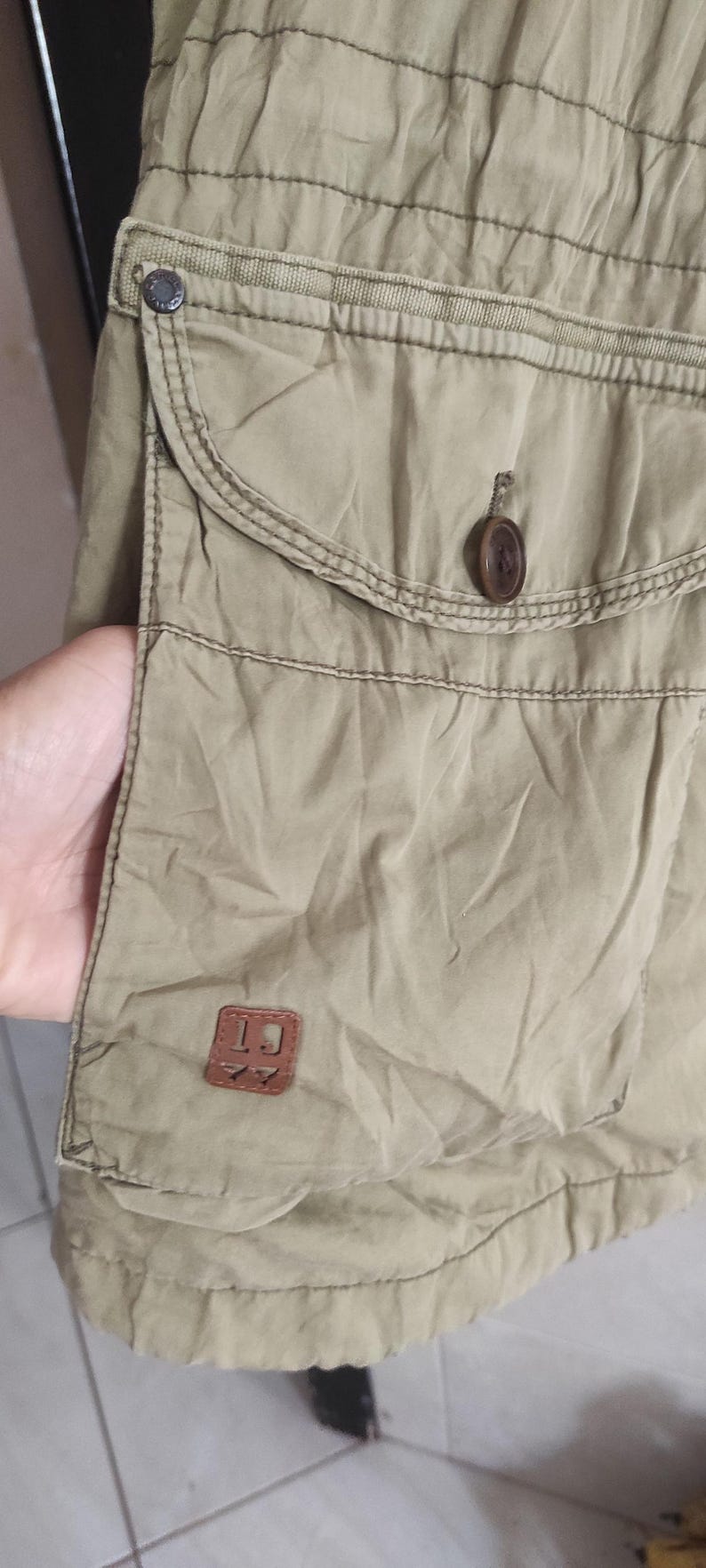May include: A khaki cargo skirt with a large pocket on the front. The pocket has a brown leather patch with the letters "LJ" on it. There is a button on the pocket flap.