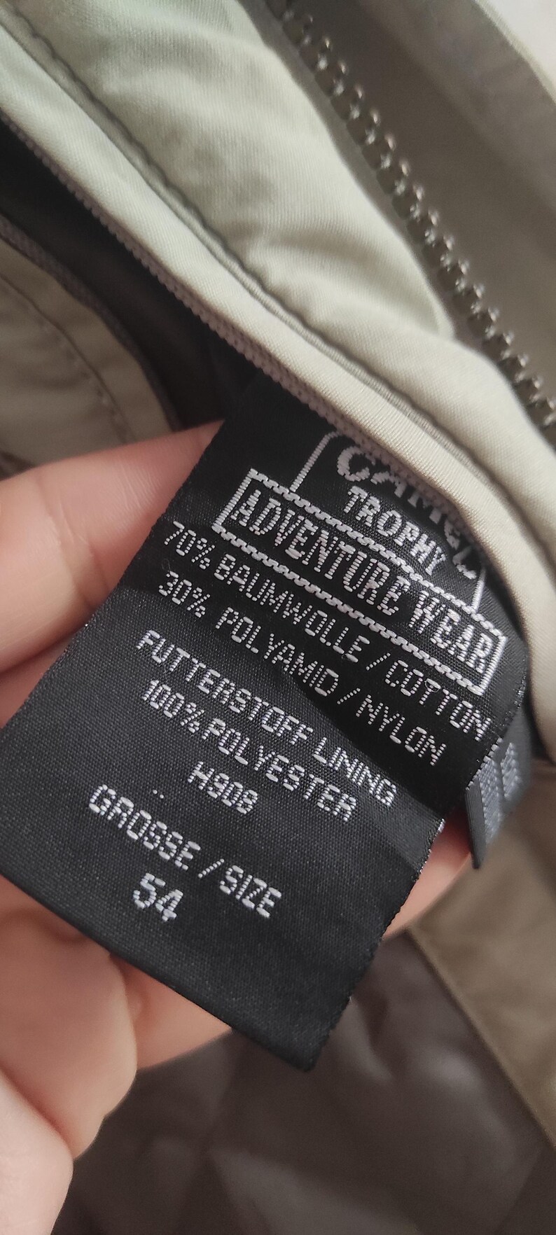 May include: A black label with white text that reads "Adventure Wear Trophy" and "70% Baumwolle/Cotton 30% Polyamid/Nylon" and "Futterstoff Lining 100% Polyester" and "H908" and "Grosse/Size 54".