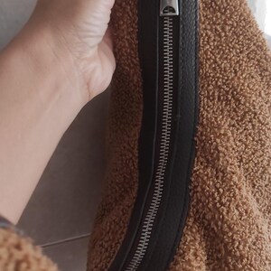May include: Brown teddy bear fabric bag with a black leather zipper and strap. The bag has a silver zipper pull. The bag is a soft, plush material.