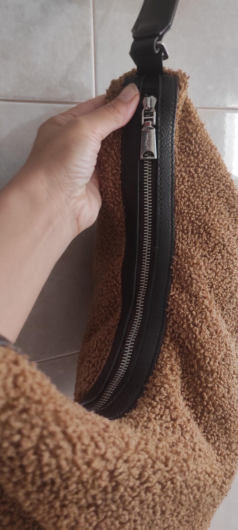 May include: Brown teddy bear fabric bag with a black leather zipper and strap. The bag has a silver zipper pull. The bag is a soft, plush material.