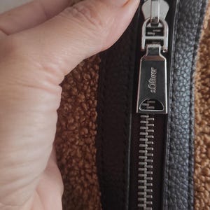 May include: Close-up of a brown and black handbag with a silver zipper. The zipper pull is engraved with the brand name "s.Oliver". The bag has a textured, faux-fur exterior.
