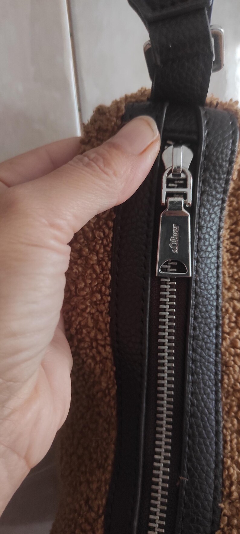 May include: Close-up of a brown and black handbag with a silver zipper. The zipper pull is engraved with the brand name "s.Oliver". The bag has a textured, faux-fur exterior.