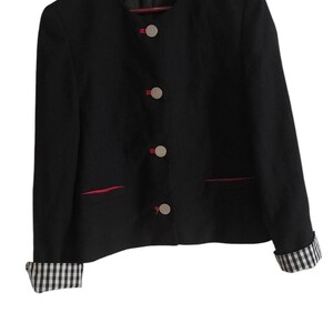 Pode incluir: Blazer preto com gola redonda, acabamentos vermelhos e botões prateados. Os punhos e o forro interior apresentam um padrão xadrez preto e branco. O casaco está pendurado num cabide de madeira.